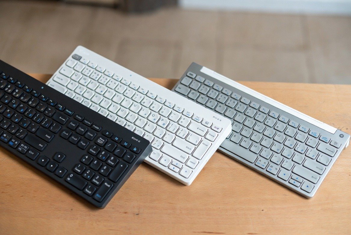 Título do post 1 Three wireless Bluetooth keyboards in black, white, and silver on a wooden desk.